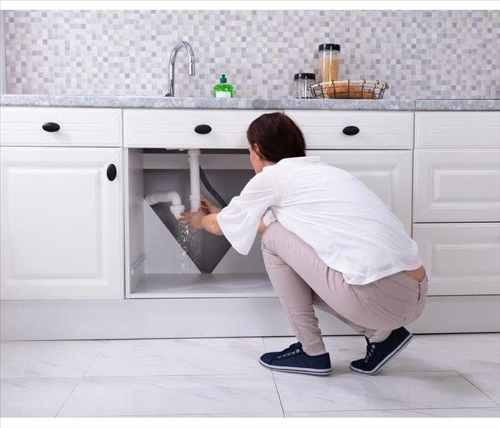 Woman in white shirt with dark hair holding pipe with water coming out under kitchen sink